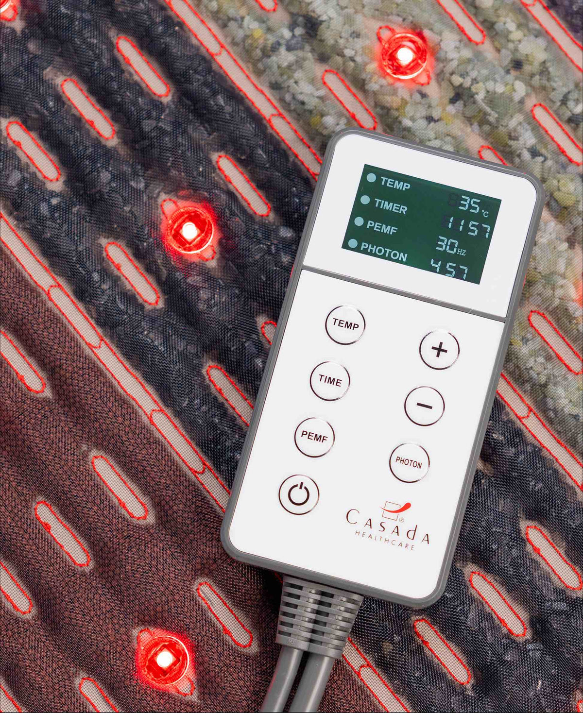 Pilot sterujący Casada z czytelnym wyświetlaczem LCD do regulacji temperatury, czasu i trybu fotonowego, położony na podgrzewanej macie masującej z czerwonymi diodami LED.